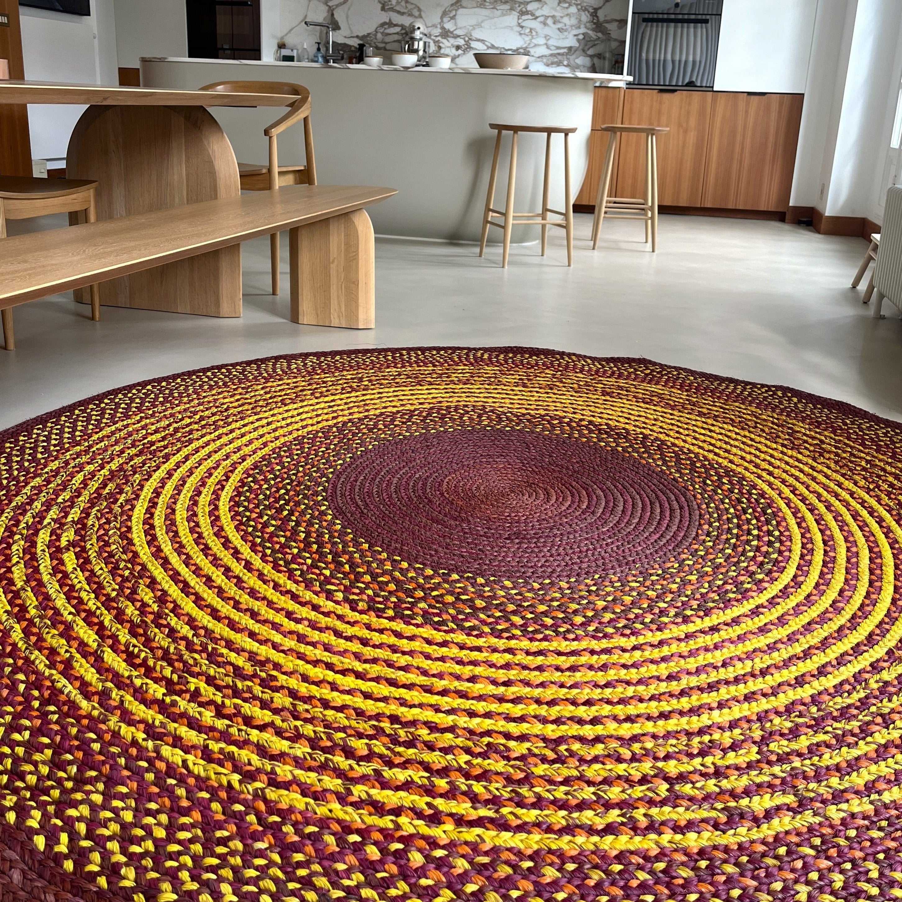 Tapis fait main en raphia avec des tons chaleureux de jaune, brun et prune, parfait pour une décoration naturelle, diamètre 230 cm.